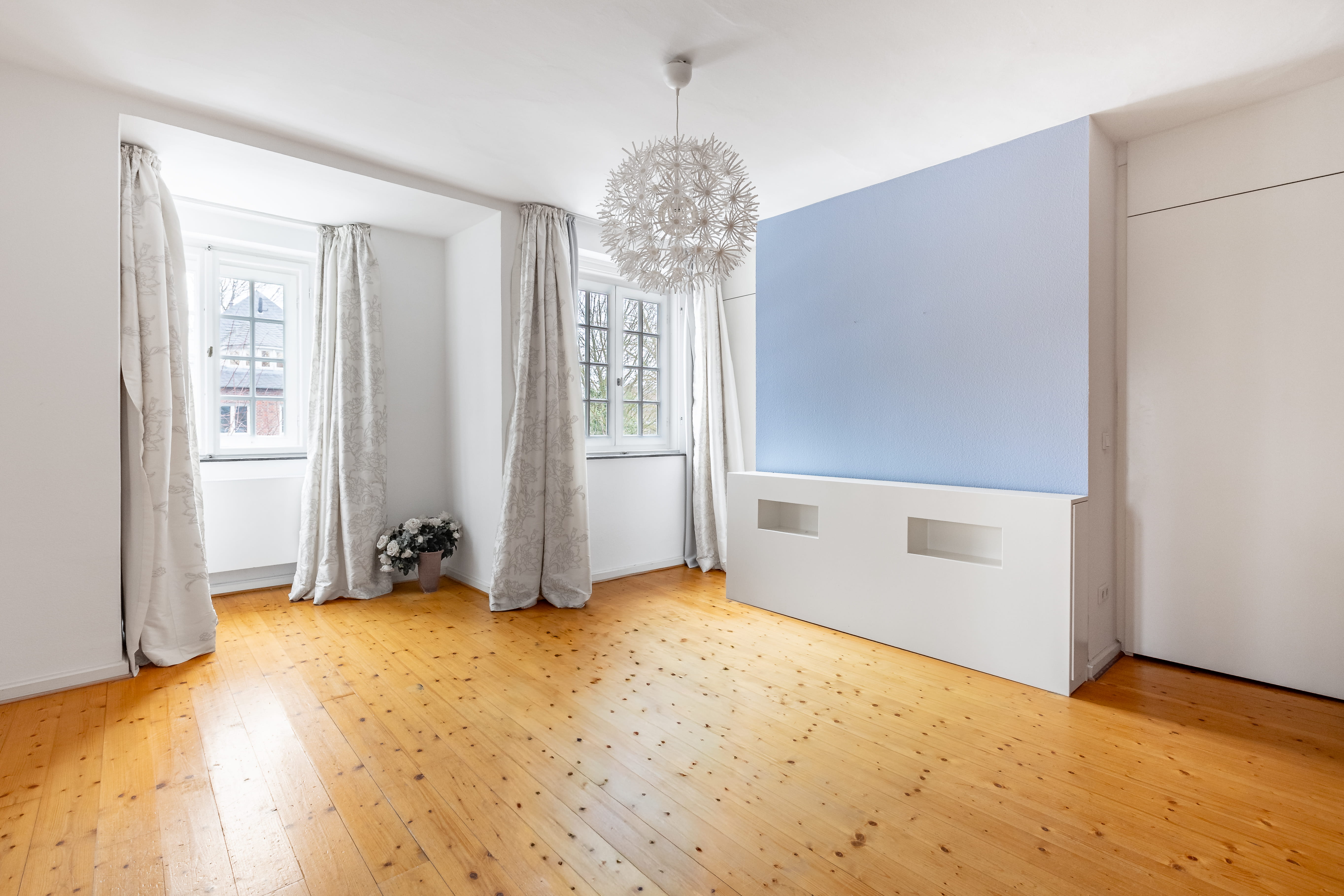 Vorher-Bild für virtuelles Home Staging: Ein leeres, unmöbliertes Schlafzimmer mit einem hellen, rustikalen Dielenboden. Der Raum verfügt über zwei Fenster mit hellen Vorhängen, eine auffällige kugelförmige Deckenleuchte und eine hellblaue Akzentwand mit einer weißen, halbhohen Vorwandinstallation. Dieser leere Zustand bietet die ideale Ausgangsbasis für eine fotorealistische digitale Möblierung, um potenziellen Käufern bei der Immobilienvermarktung die optimale Raumnutzung und das volle Potenzial zu veranschaulichen.